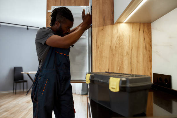 Side view concerned African American service man in workwear looking inside opened refrigerator, lighting with flashlight. Toolkit placed near on kitchen surface. Modern appartment on background.