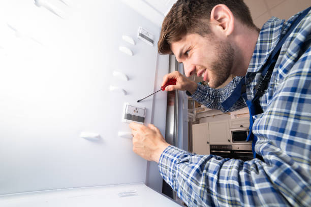 Side view concerned African American service man in workwear looking inside opened refrigerator, lighting with flashlight. Toolkit placed near on kitchen surface. Modern appartment on background.