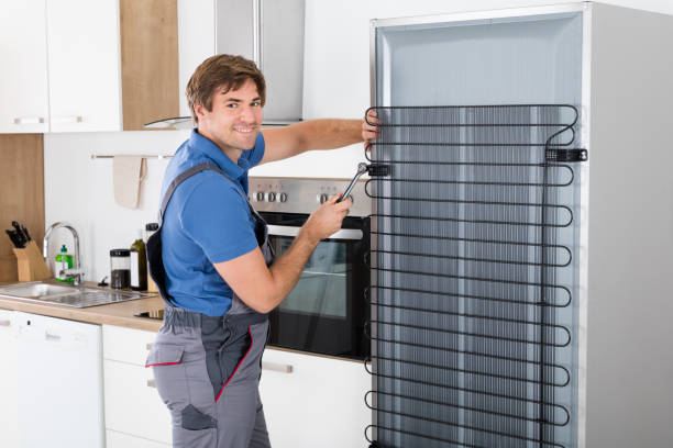 Serviceman In Overall Working On Fridge With Screwdriver In Kitchen